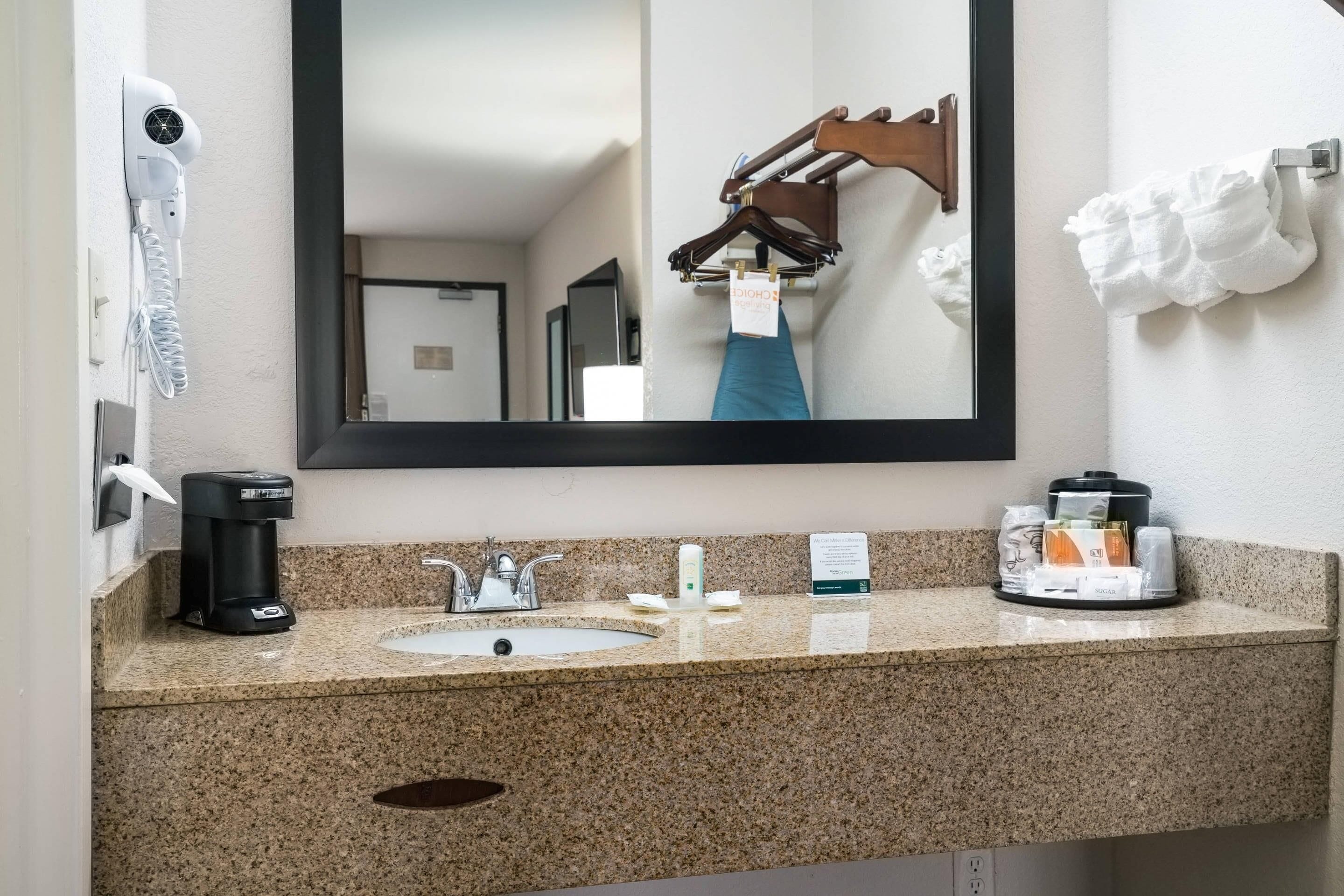 Un baño de hotel con encimera de granito, un lavabo, un espejo, un perchero para toallas, un secador de pelo y una cafetera.