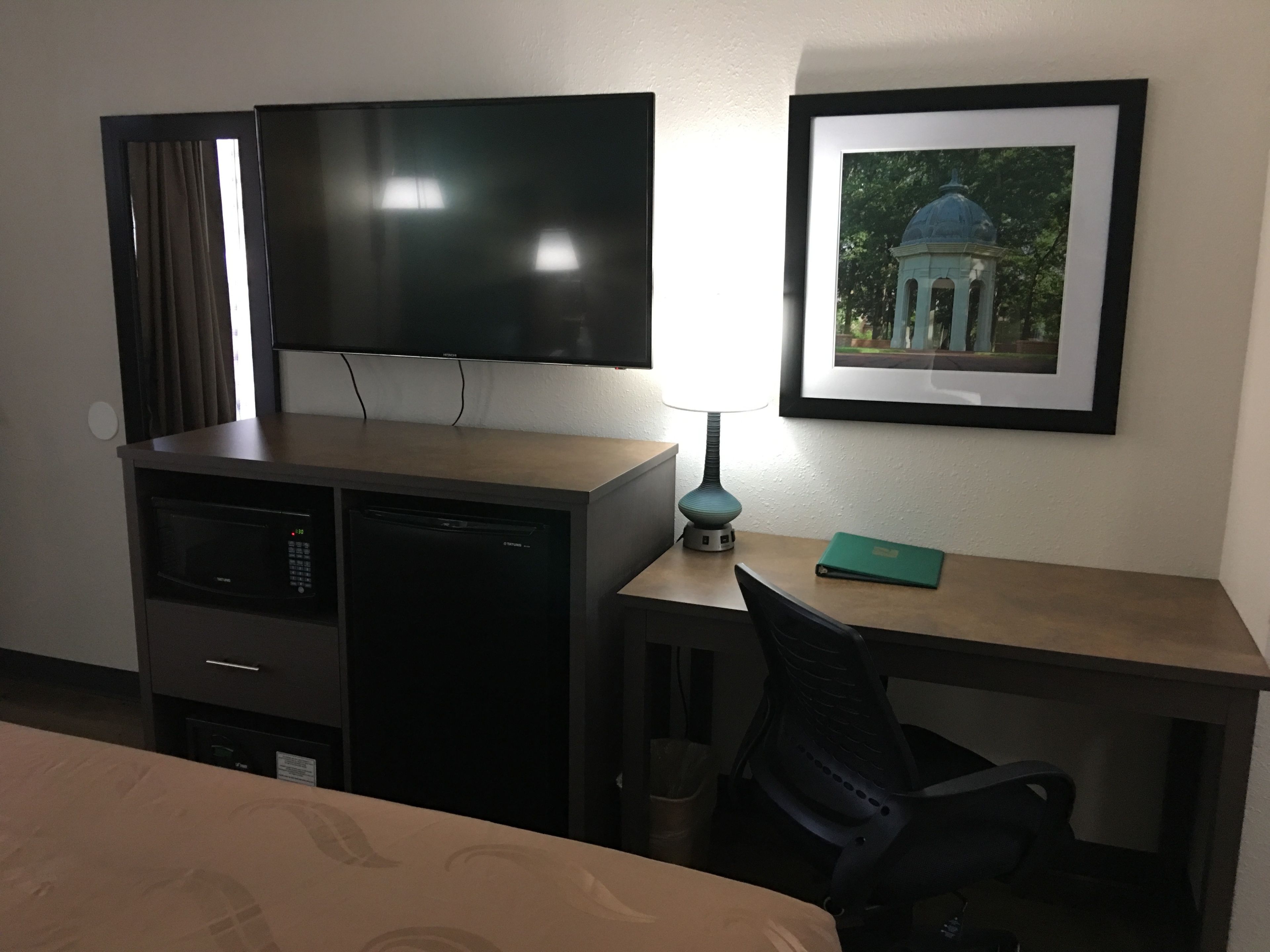 A hotel room with a bed, a desk with a chair, a television, a framed picture on the wall, and a lamp.