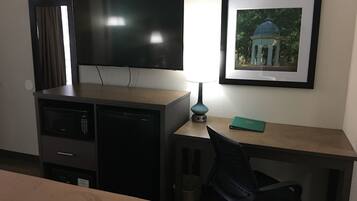 A hotel room with a bed, a desk with a chair, a television, a framed picture on the wall, and a lamp.