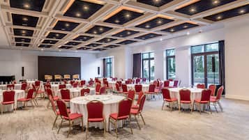 A large conference room with round tables and red chairs, a high ceiling with recessed lighting, and large windows providing natural light.