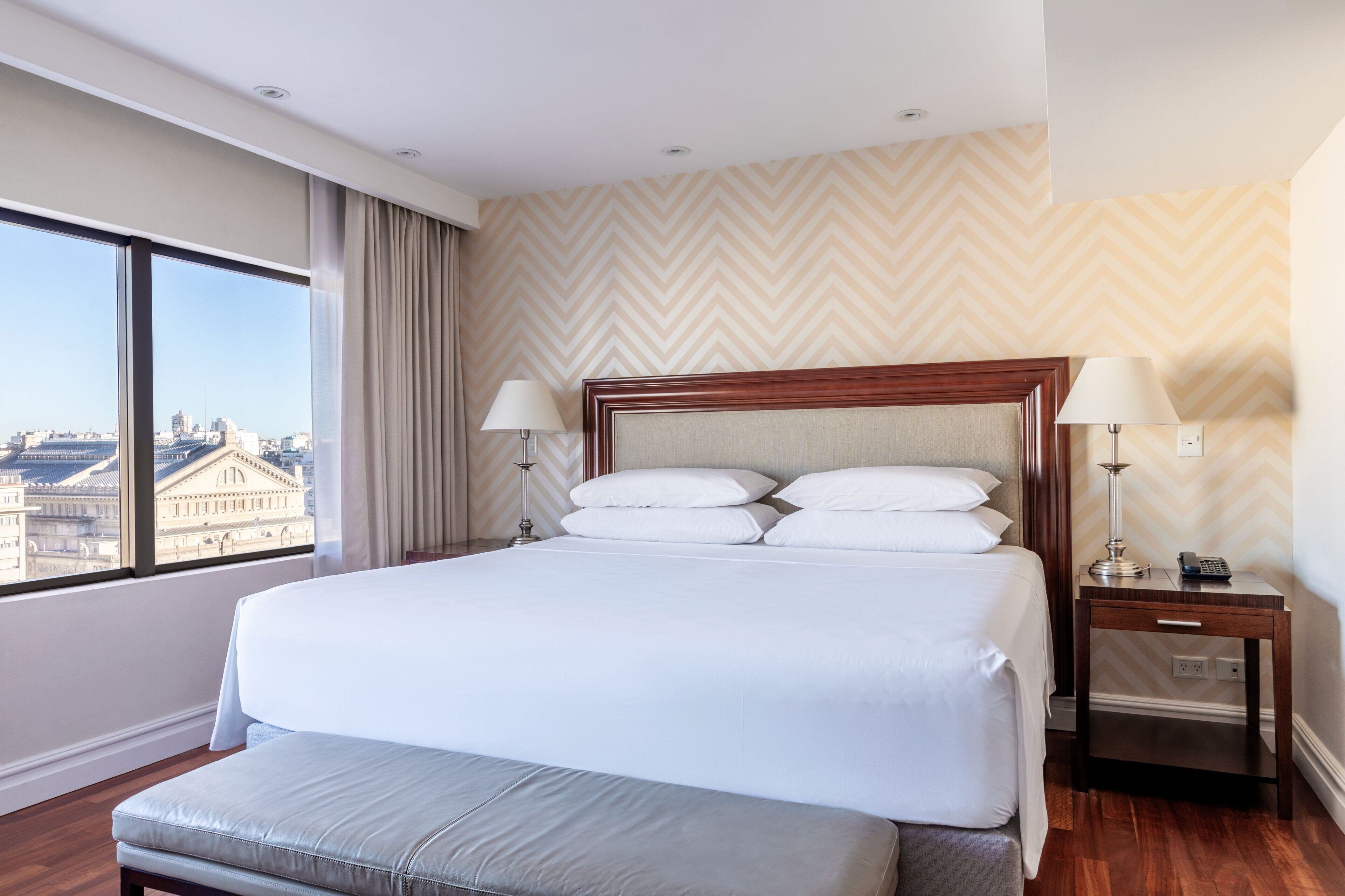 Hotelkamer met een groot bed, twee bedlampen, een houten nachtkastje en uitzicht op een stadsgezicht door grote ramen.