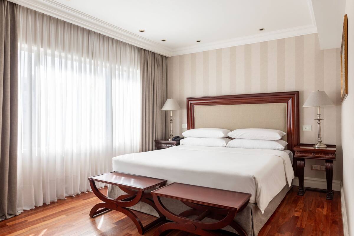 A hotel room with a large bed, two bedside tables, a nightstand, and a window with curtains.