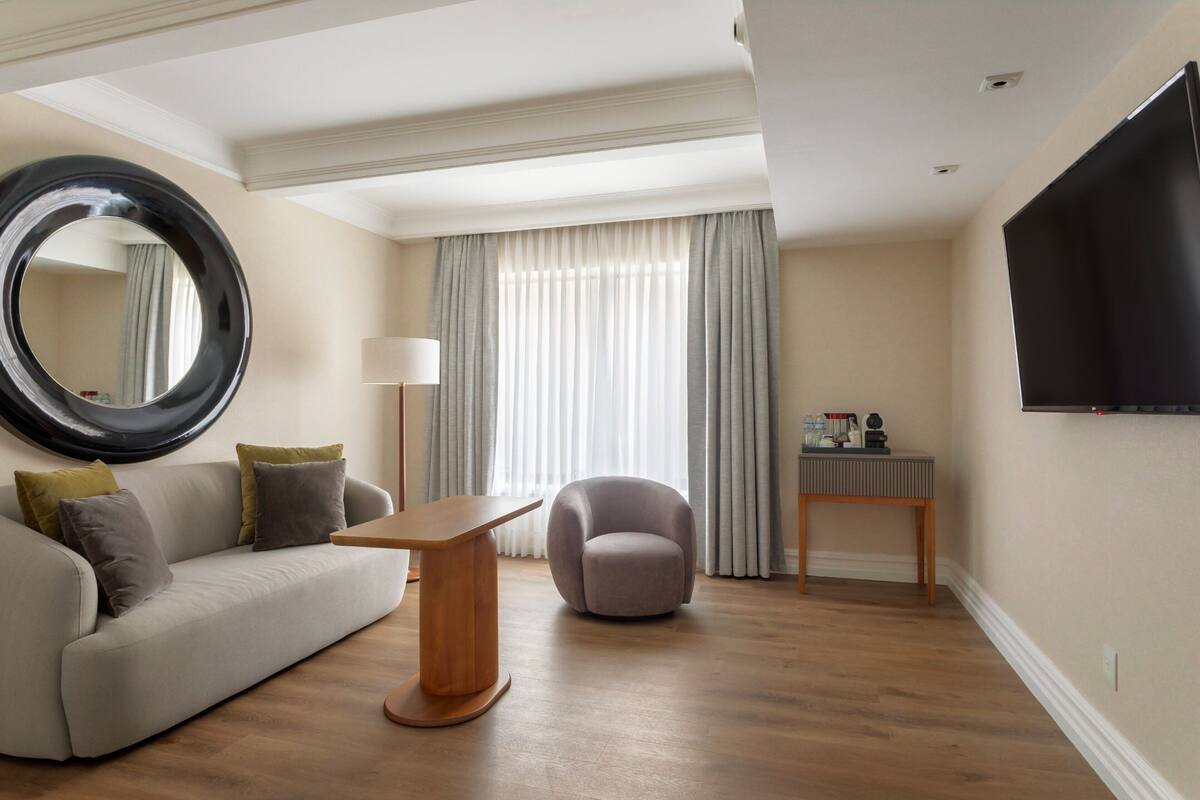 A modern living room with a sofa, a round mirror, a small table, a round chair, a TV, and a window with curtains.