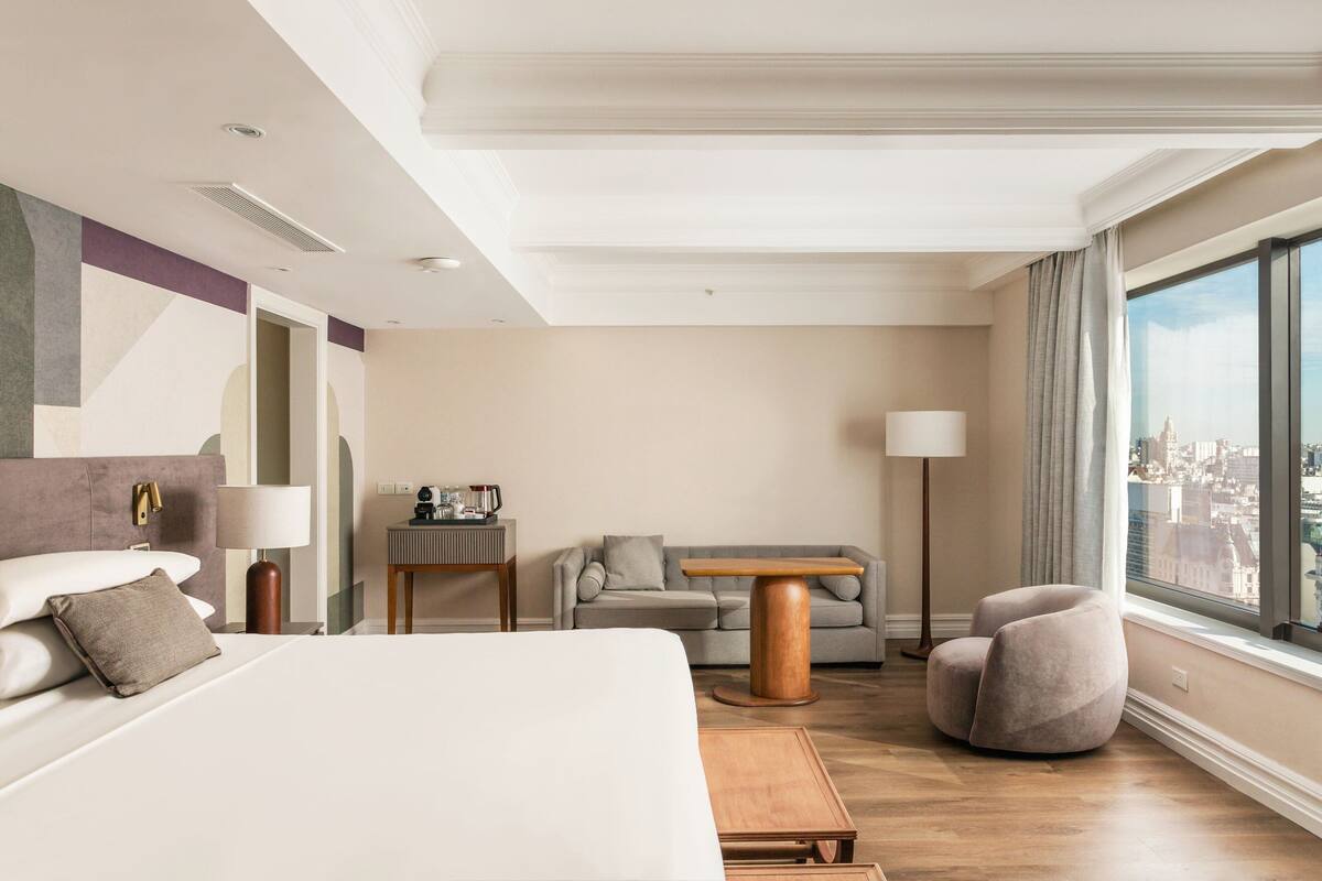 A modern hotel room with a large bed, a sofa, a small table, and a chair. There is a city view through the window.