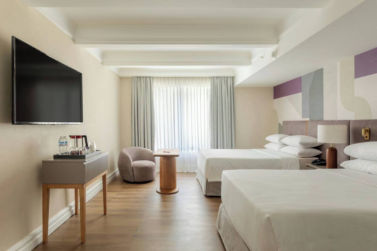 A hotel room with a flat-screen TV, a desk with toiletries, two beds, a chair, and a large window with curtains.