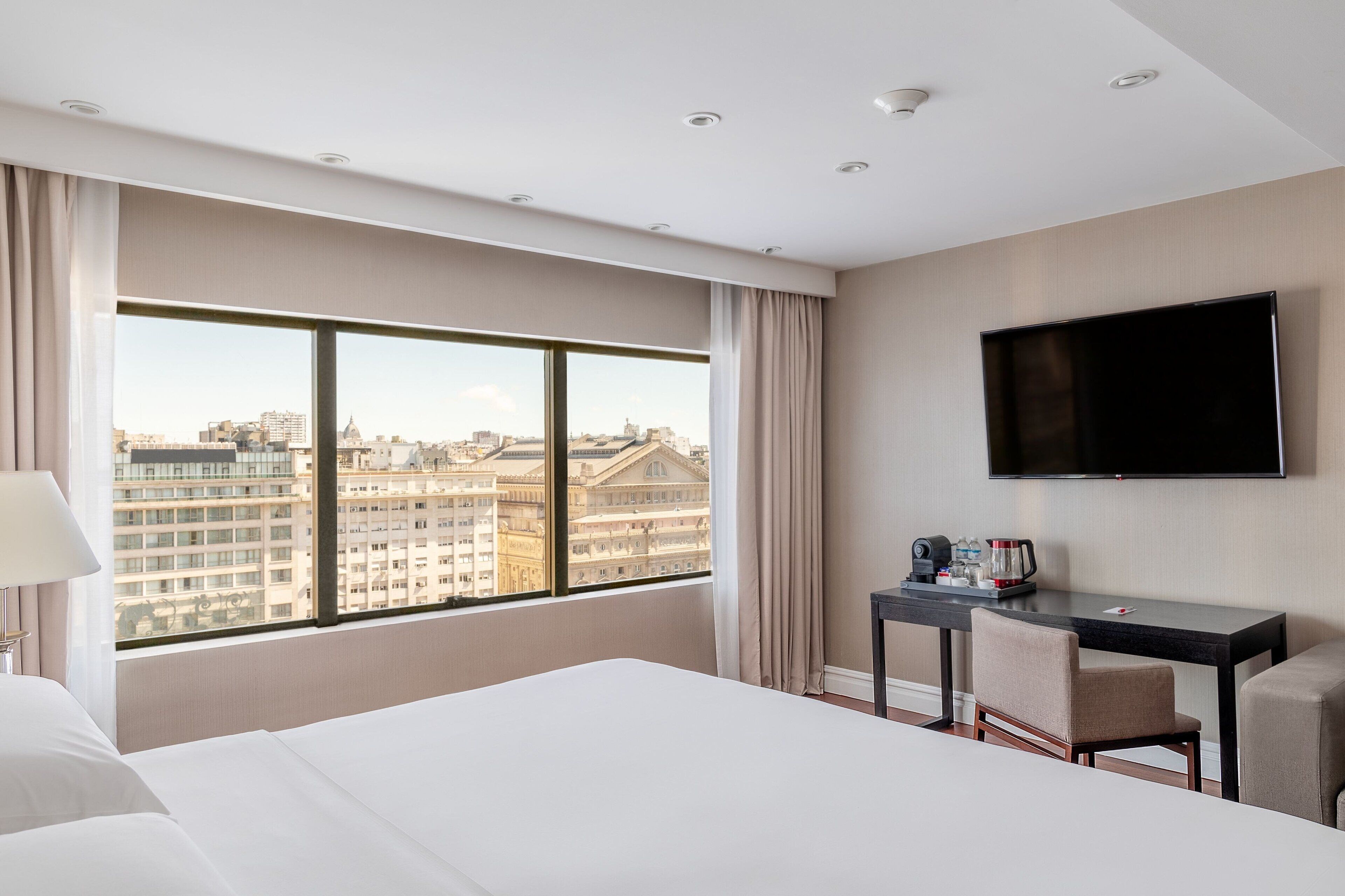 A hotel room with a large window, a bed, a desk with a chair, and a flat-screen TV on the wall.