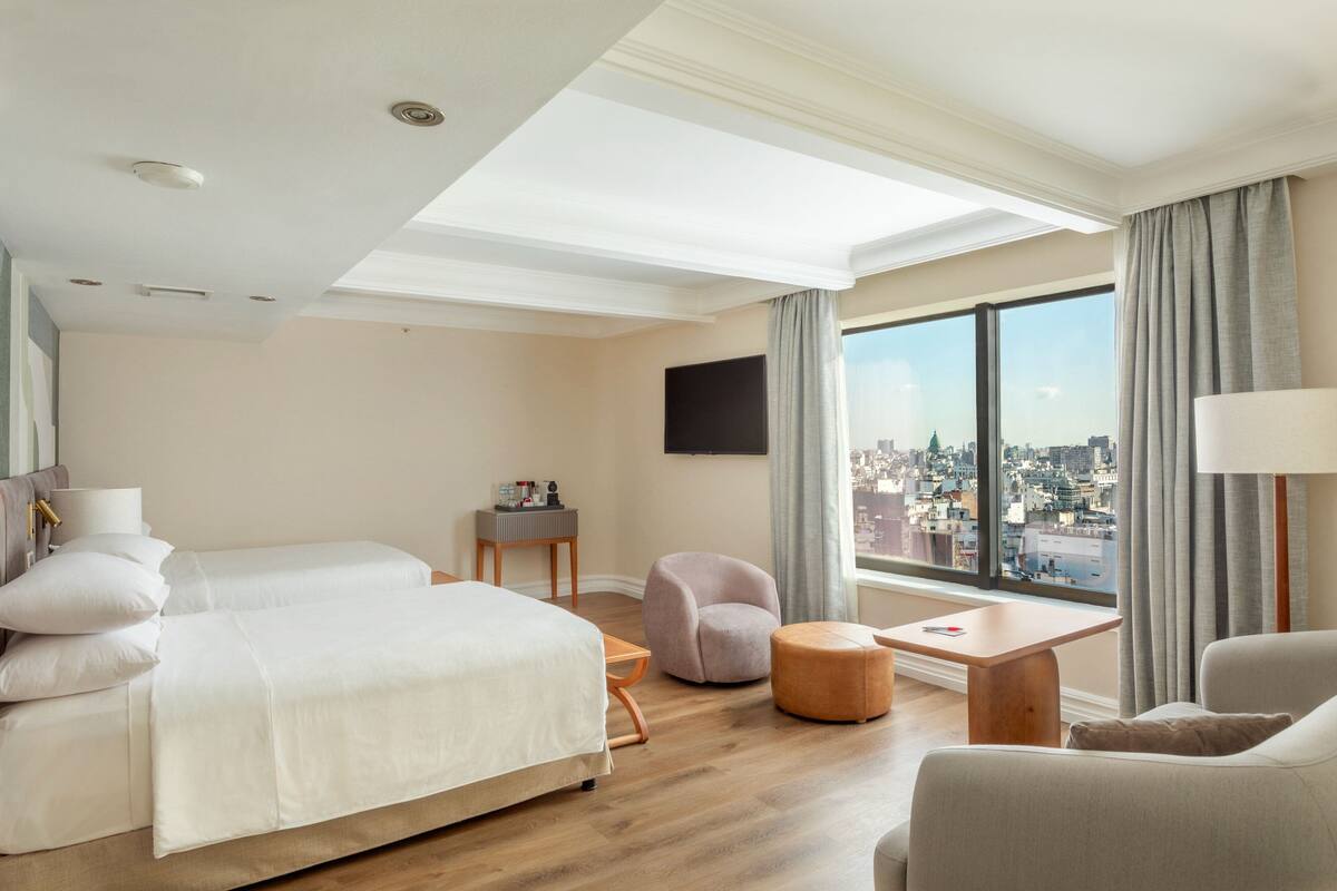 A hotel room with a bed, a seating area, a TV, and a large window with a city view.