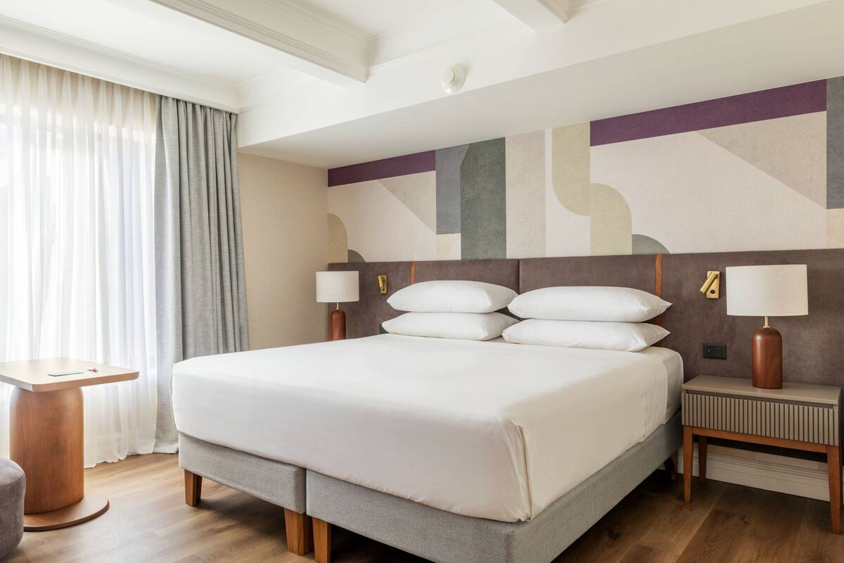 A hotel room with a large bed, a bedside table, a nightstand, a wall with abstract art, and a window with curtains.