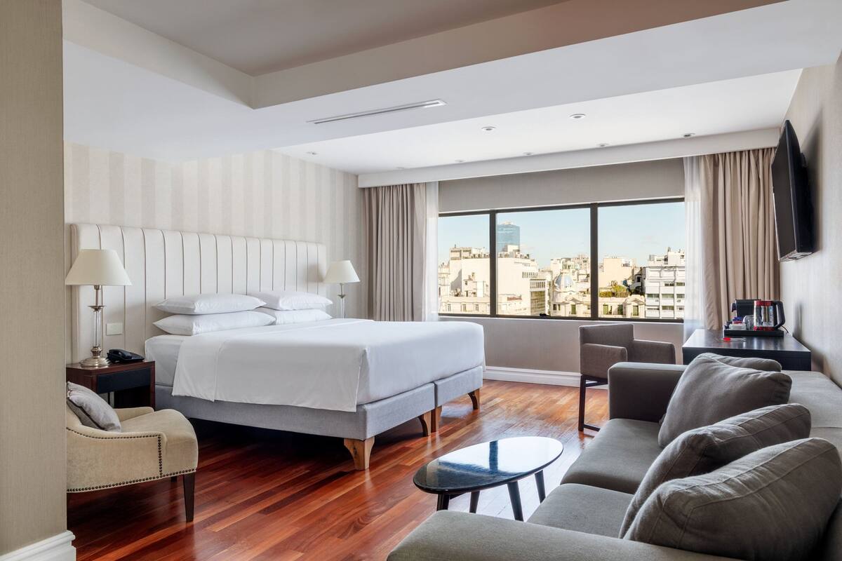 A modern hotel room with a large bed, a sofa, a small table, and a city view through the window.