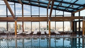 Piscina interna con piastrelle a scacchi, poltrone relax e vista sulla città attraverso ampie finestre.