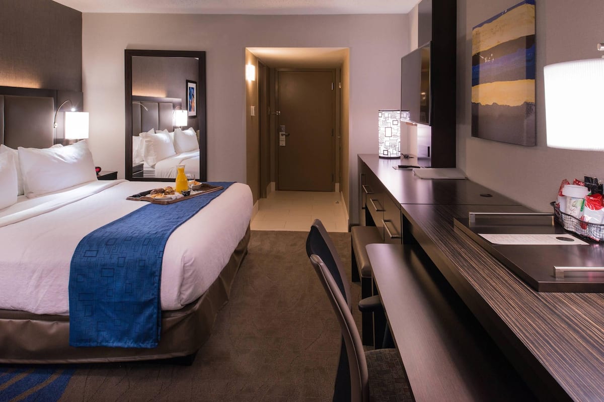 A hotel room with a bed, a desk, a chair, a mirror, a TV, and a bathroom.