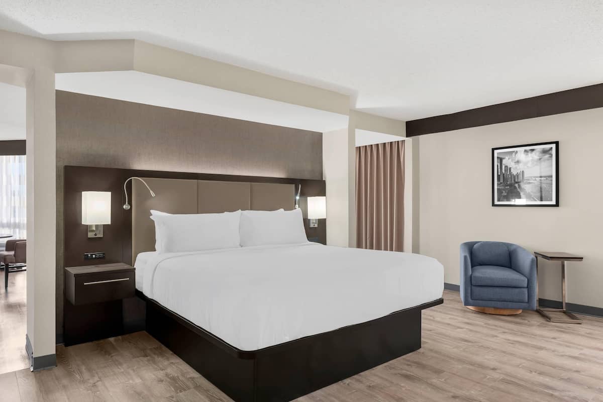 A modern hotel room with a large bed, bedside lamps, a chair, and a framed picture on the wall.