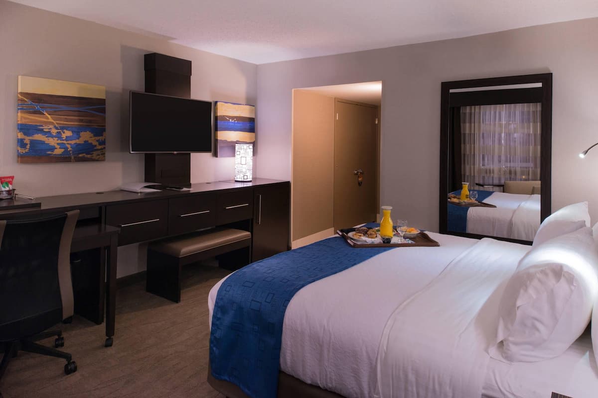 A hotel room with a bed, desk, chair, television, and artwork on the wall.