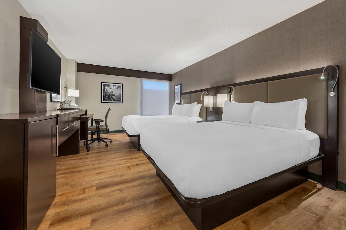 A hotel room with two beds, a desk, a flat-screen TV, and a large window.