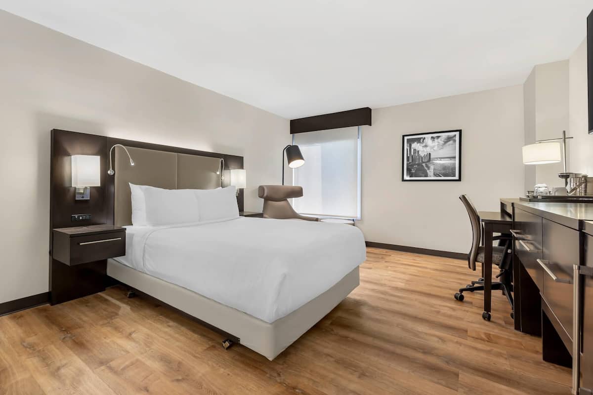 A modern hotel room with a large bed, bedside tables, a desk with a chair, and a window.