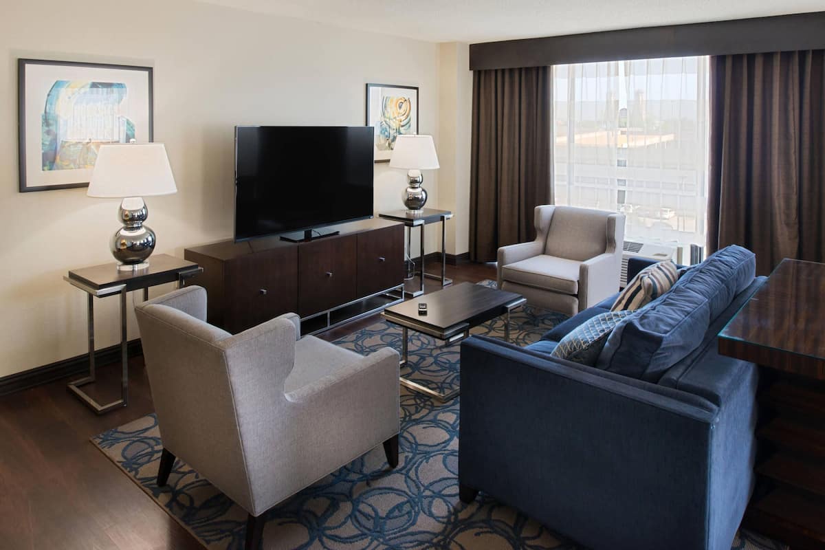 A hotel room with a flat-screen TV, two armchairs, a sofa, a coffee table, and a small side table with a lamp.