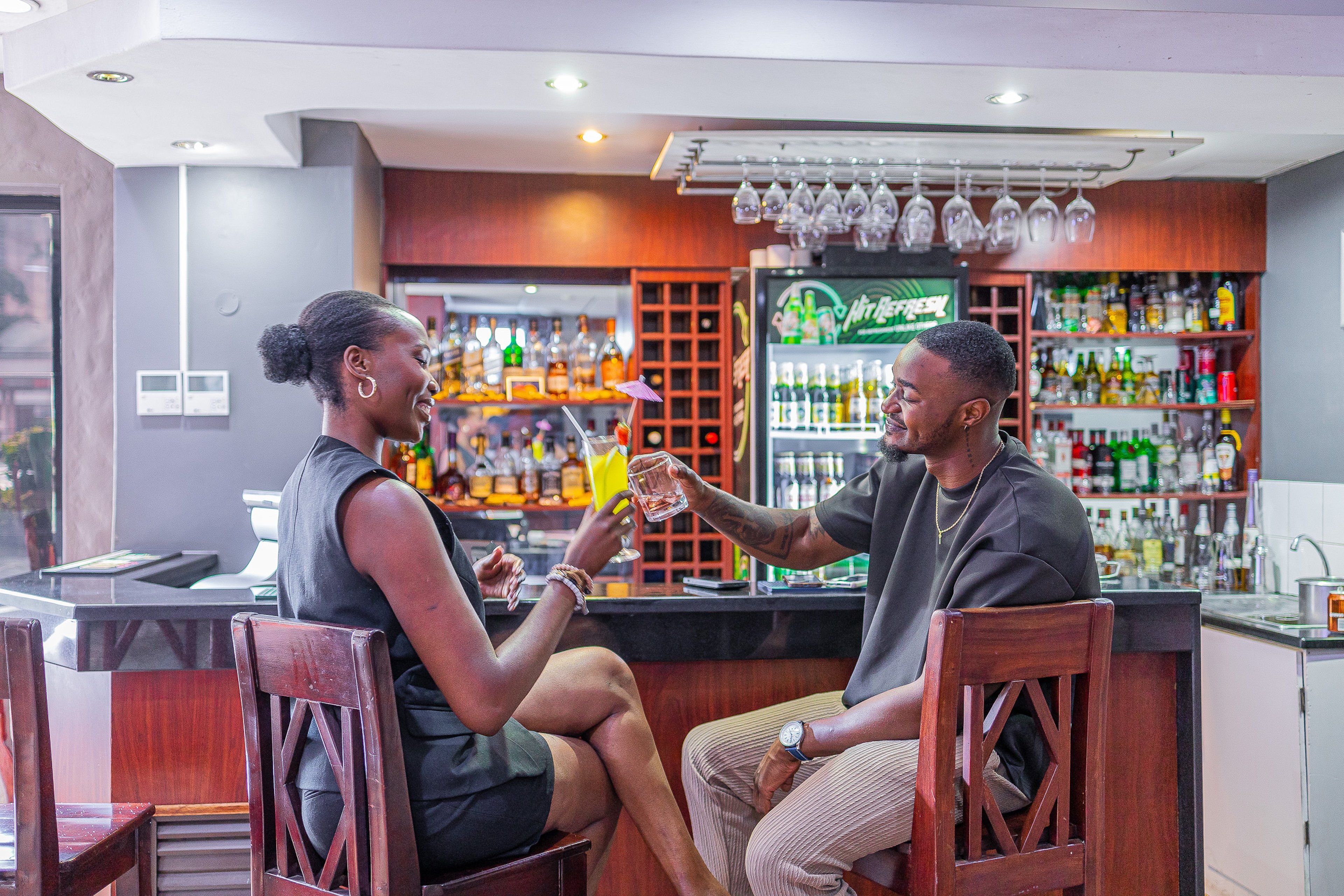 Two people seated at a bar with a variety of bottles and glasses in the background.