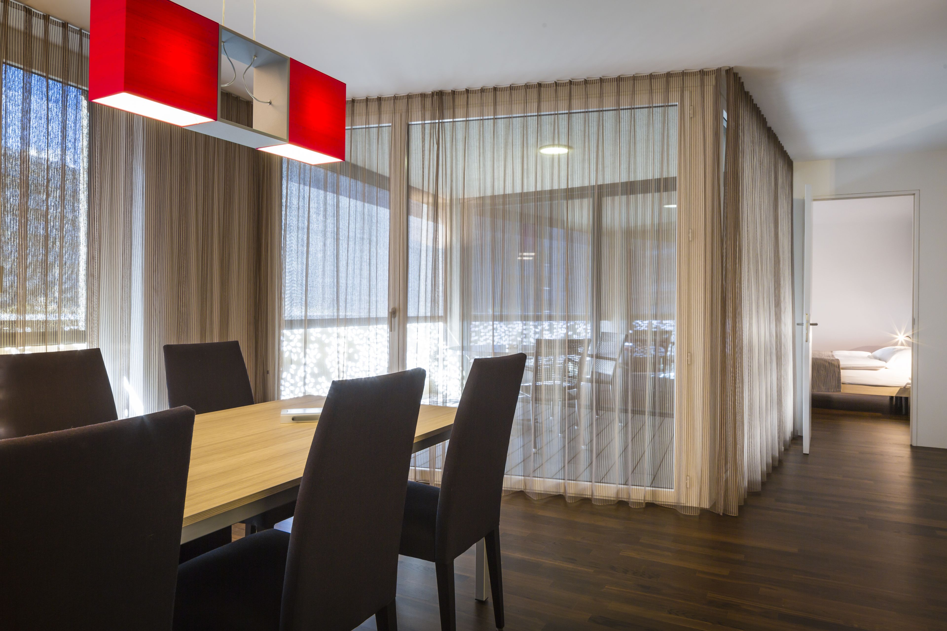 Un espace de repas moderne avec une table et des chaises en bois, de grandes fenêtres ornées de rideaux légers et une vue sur l’extérieur.