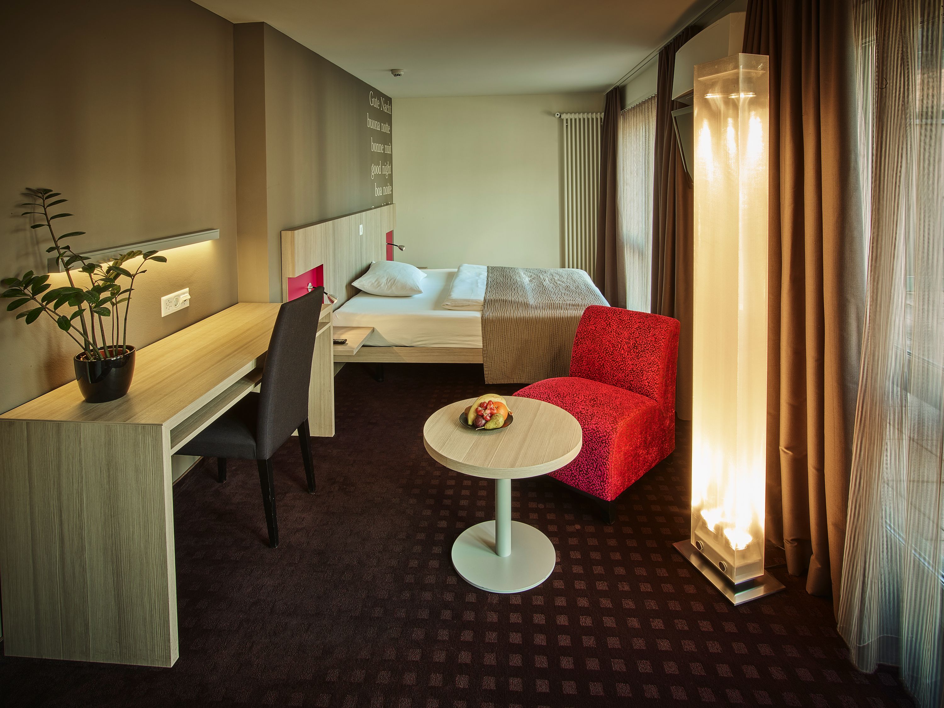 A hotel room with a bed, a desk, a chair, a small table with fruit, and a potted plant.