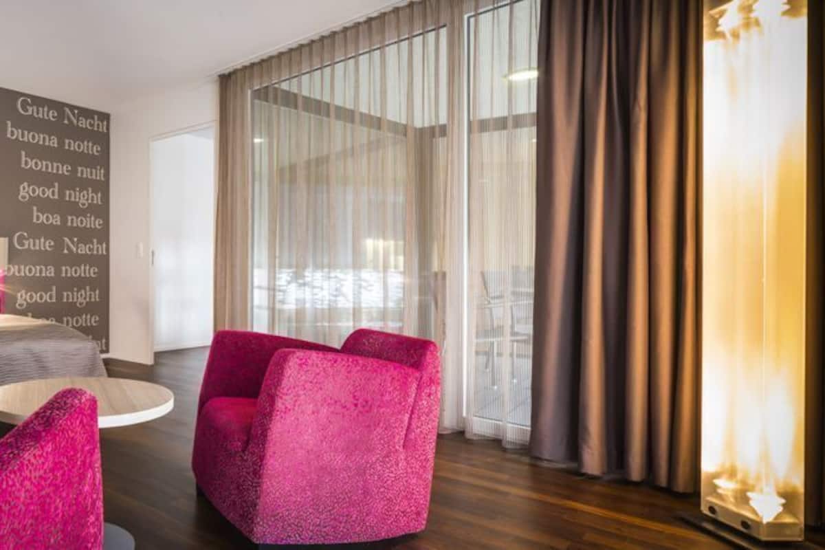 A modern hotel room with a bed, two pink armchairs, a round coffee table, and a large window with curtains.
