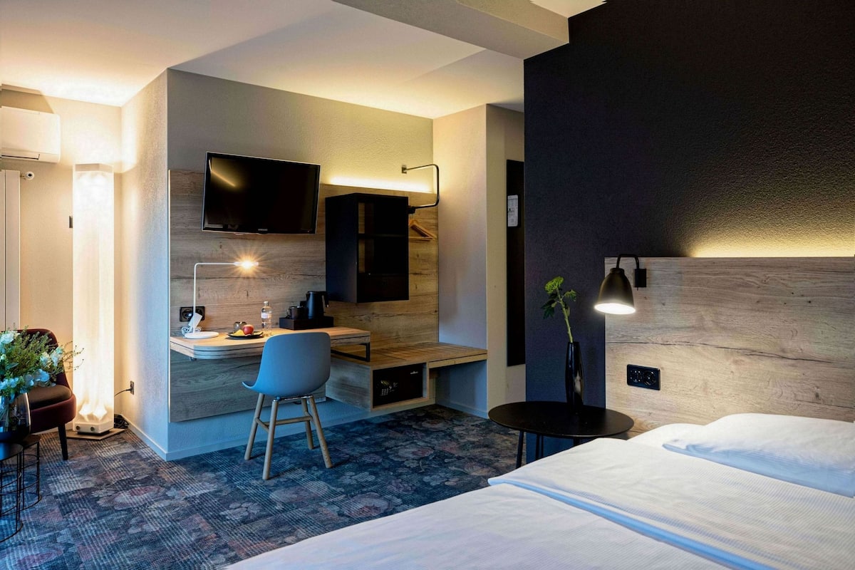 A modern hotel room with a bed, a desk with a chair, a television, and a small table with a plant.