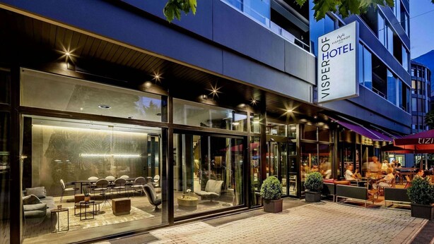 A modern hotel exterior with a glass facade, outdoor seating area, and a sign indicating it is the Visperhof Hotel. - Baxter Hotel (Visp)