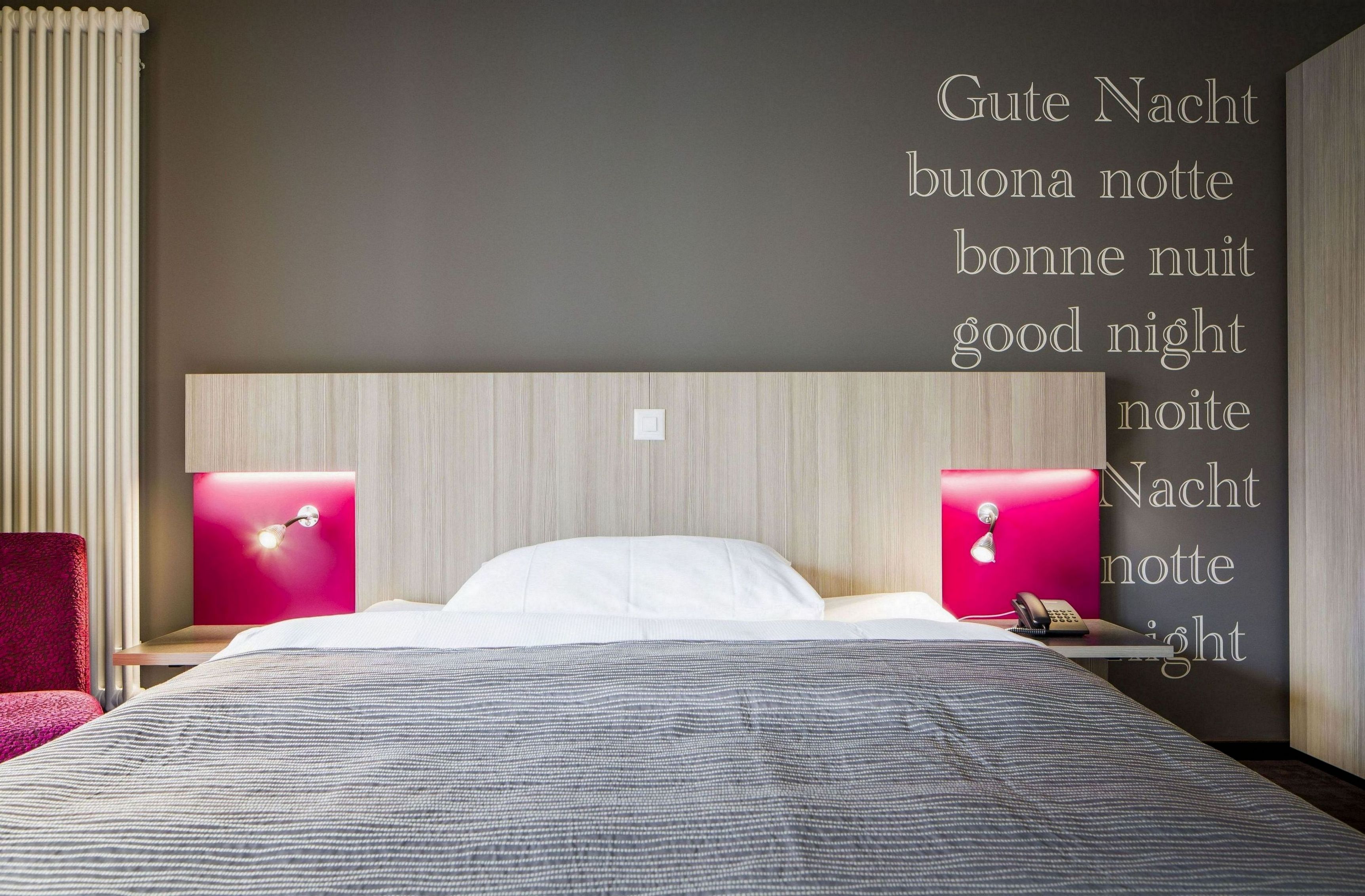 Et hotelværelse med en seng, to sengeborde med lamper, en rød stol og en væg med tekst på flere sprog, der siger "Good Night".
