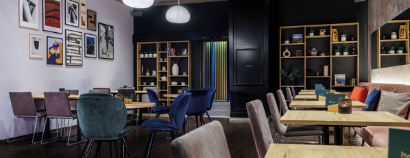 A modern cafe with wooden tables and chairs, art on the walls, and shelves with books and plants. - Baxter Hotel (Visp)