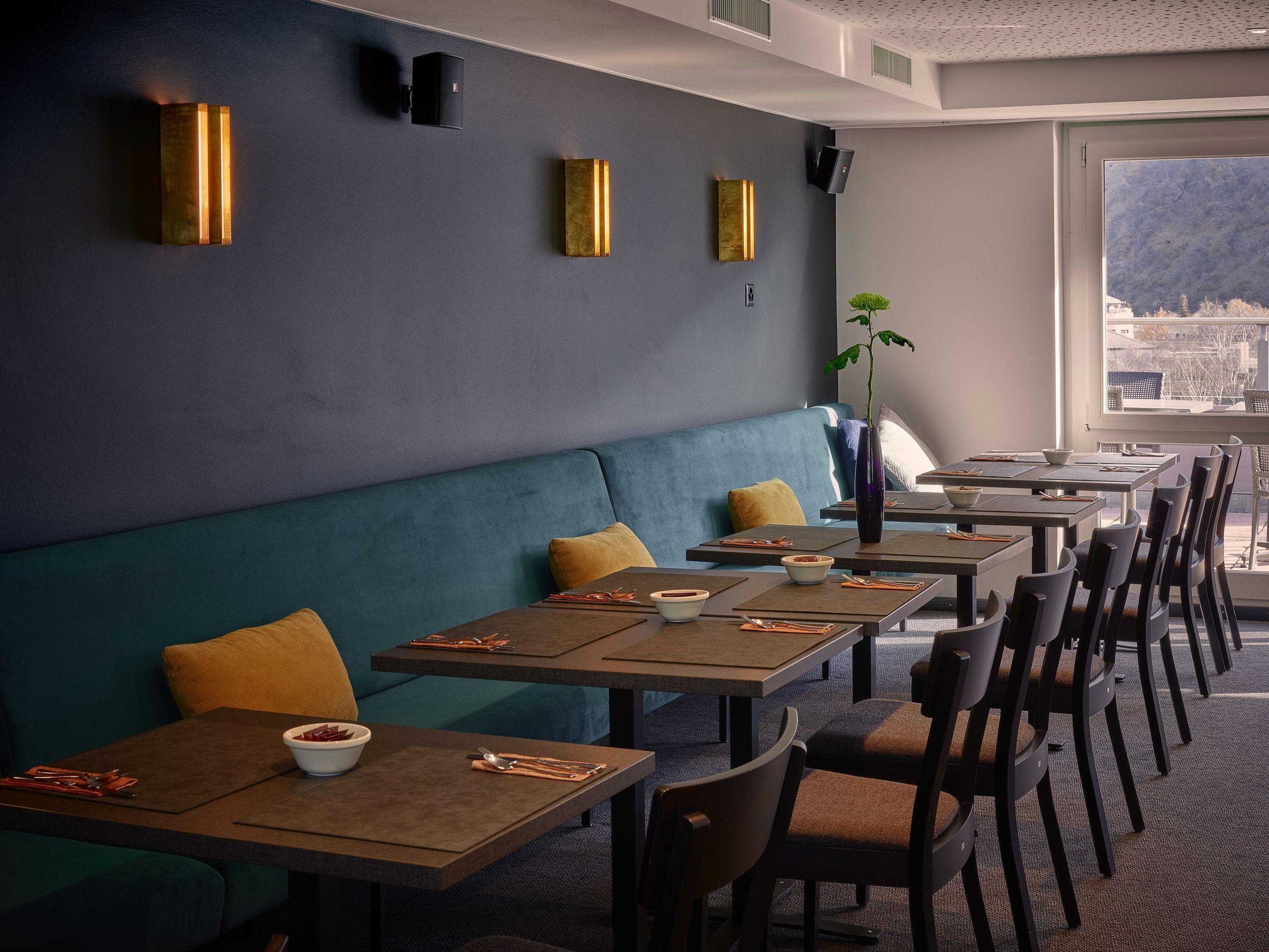 Un espace de restauration moderne avec une rangée de tables et de banquettes, une grande fenêtre donnant sur un paysage urbain et un luminaire fixé au mur.