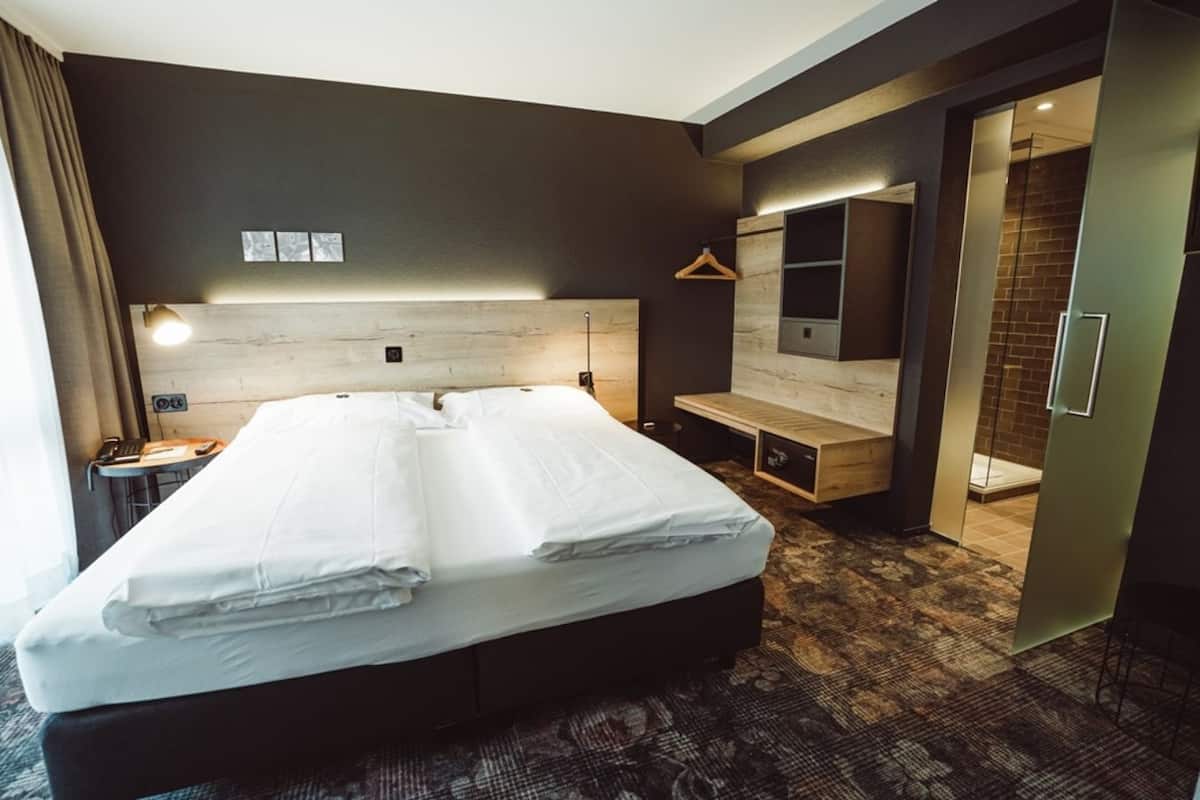 A modern hotel room with a large bed, a desk, a chair, a hanging light, and a mirror.