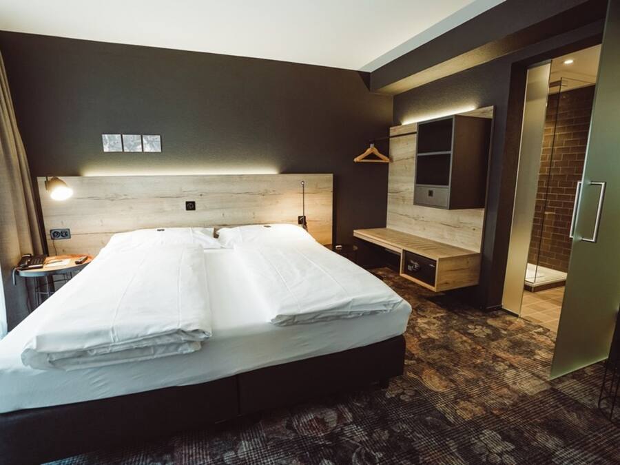 A modern hotel room with a large bed, a desk, a chair, a hanging light, and a mirror.