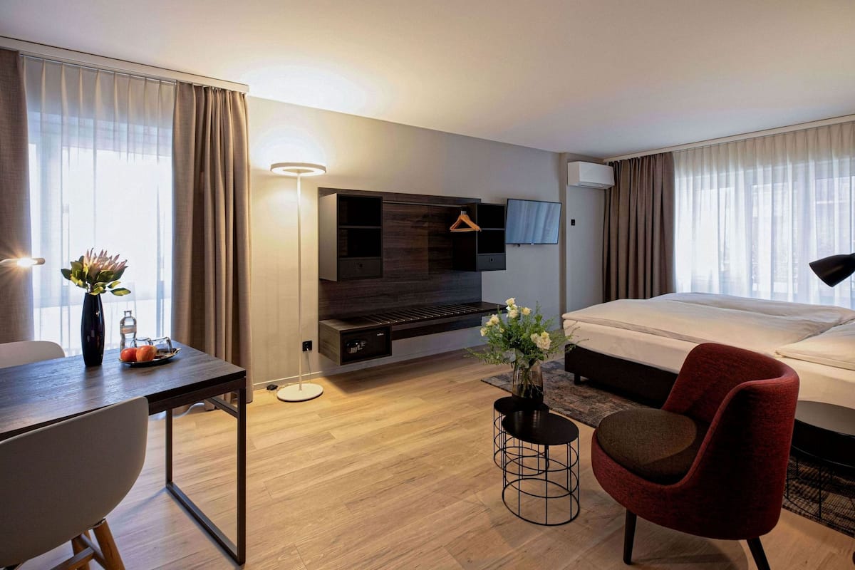 A hotel room with a bed, a desk, a chair, a TV, and a small table with a vase.