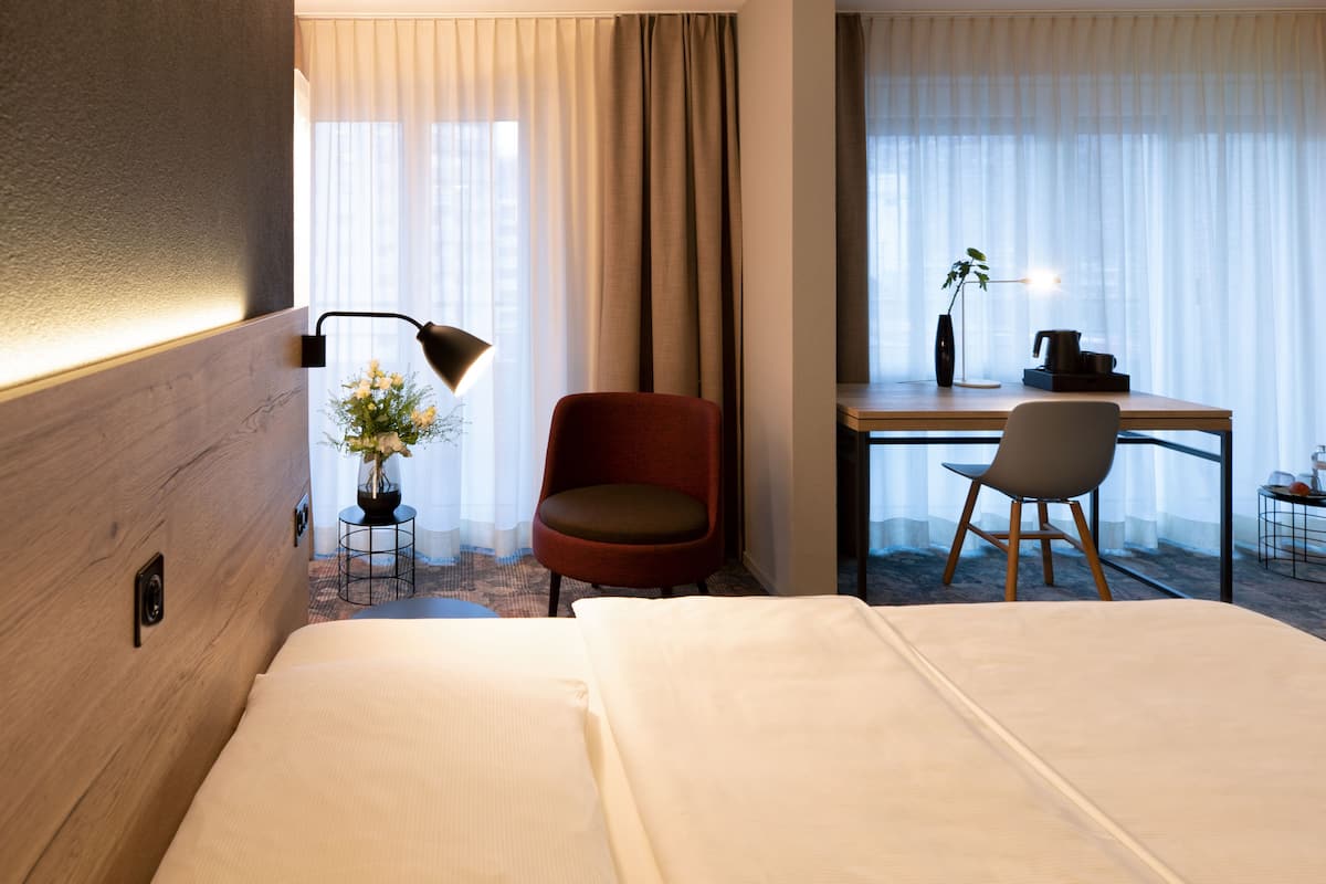 A hotel room with a bed, a desk, two chairs, a lamp, and a vase with flowers.