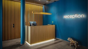 A reception area with a wooden reception desk, a blue wall with a neon 'reception' sign, and a small dog statue. - Baxter Hotel (Visp)