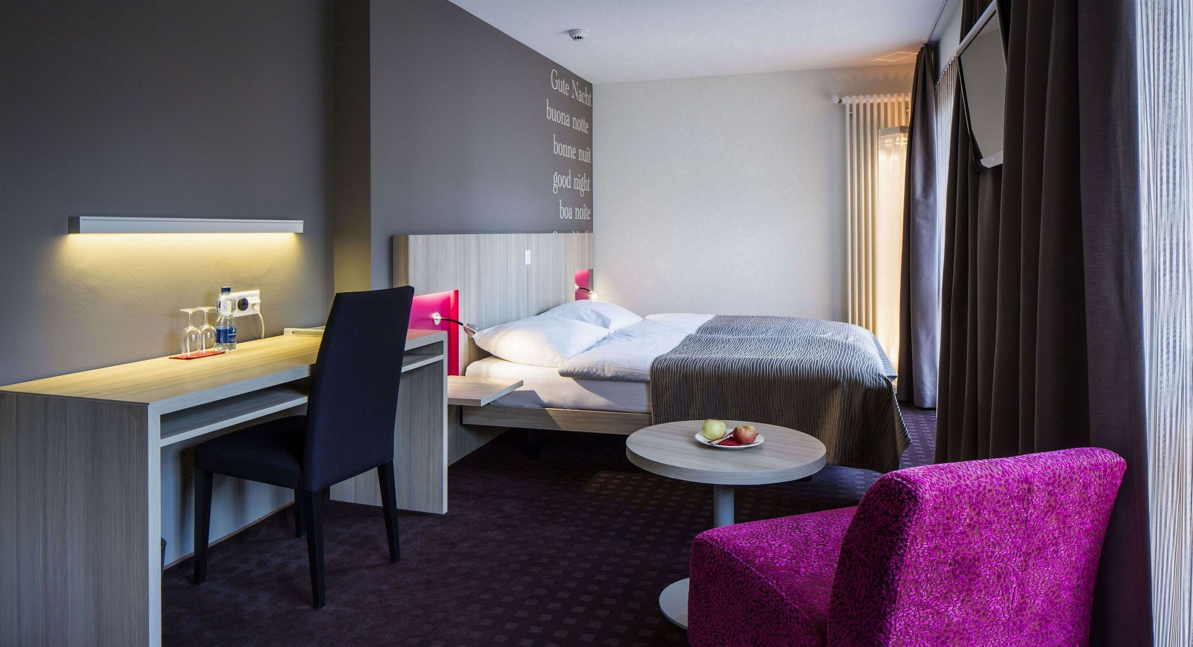 A hotel room with a bed, a desk with a chair, a small round table with fruit, and a pink chair.