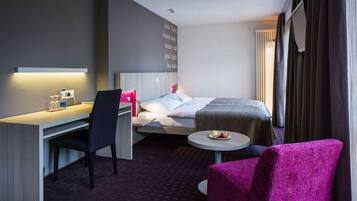 A hotel room with a bed, a desk with a chair, a small round table with fruit, and a pink chair.