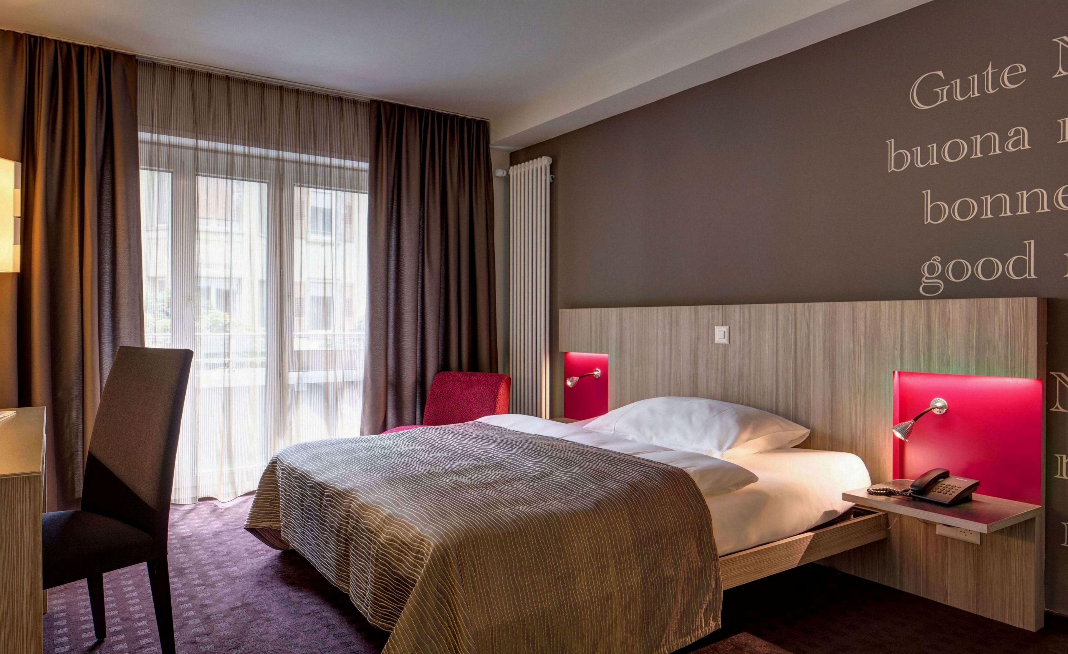 A hotel room with a bed, a desk, a chair, a red lamp, and a wall with text.