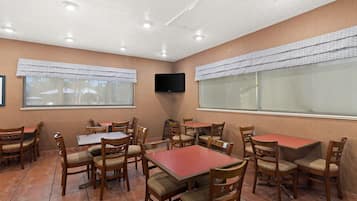 A dining area with wooden tables and chairs, a flat-screen TV mounted on the wall, and large windows with patterned curtains.
