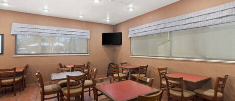 A dining area with wooden tables and chairs, a flat-screen TV mounted on the wall, and large windows with patterned curtains.