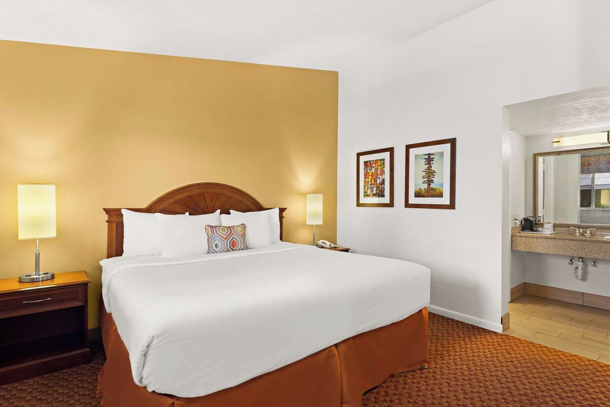 A hotel room with a large bed, bedside lamps, a nightstand, a wooden headboard, a wall-mounted telephone, and a bathroom with a sink and mirror.