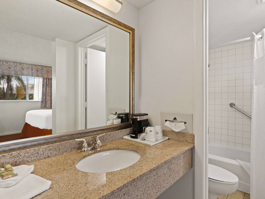 A hotel bathroom with a granite countertop, a sink, a mirror, a coffee maker, and a shower area.