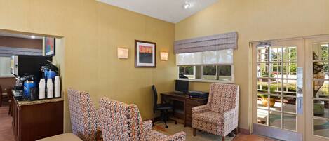 A hotel room with a kitchenette, two patterned armchairs, a desk with a computer, and a large window with a view of the outdoors.