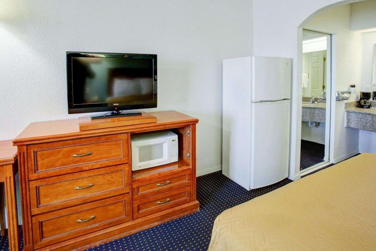 A hotel room with a wooden dresser, a television, a microwave, a refrigerator, and a bathroom with a sink and mirror.