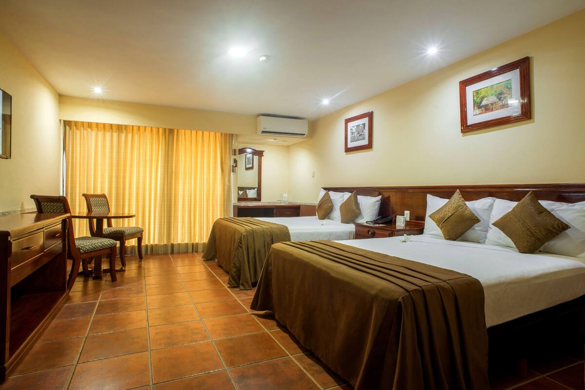 A hotel room with two beds, a desk with a chair, a small table, and a window with curtains.