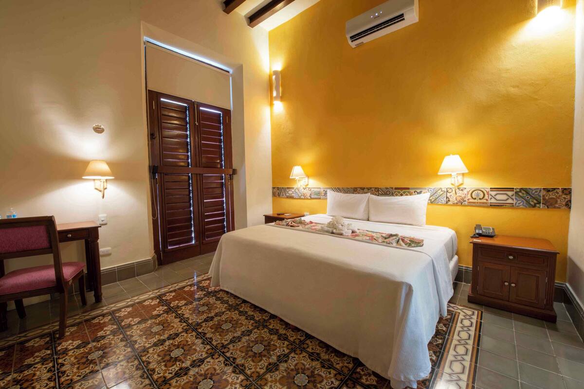 A hotel room with a bed, two bedside lamps, a wooden nightstand, a chair, and a patterned rug.