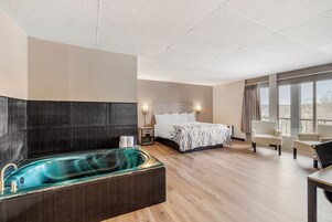 Habitación de hotel con una bañera grande, una cama con colcha estampada, una mesita de noche, un escritorio y vistas al exterior a través de amplias ventanas.