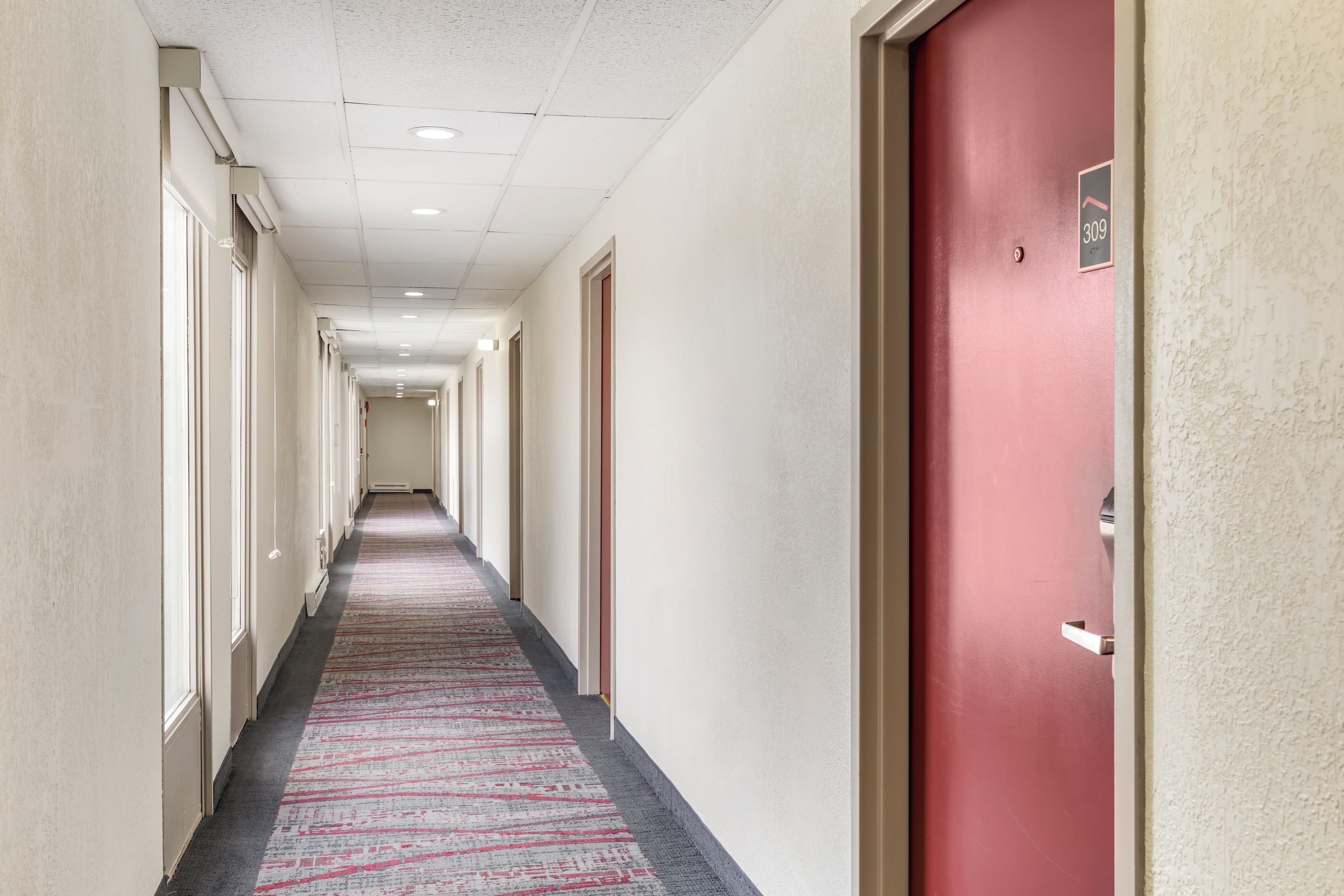 Un pasillo de hotel con alfombra estampada, puertas a ambos lados y una puerta roja a la derecha.
