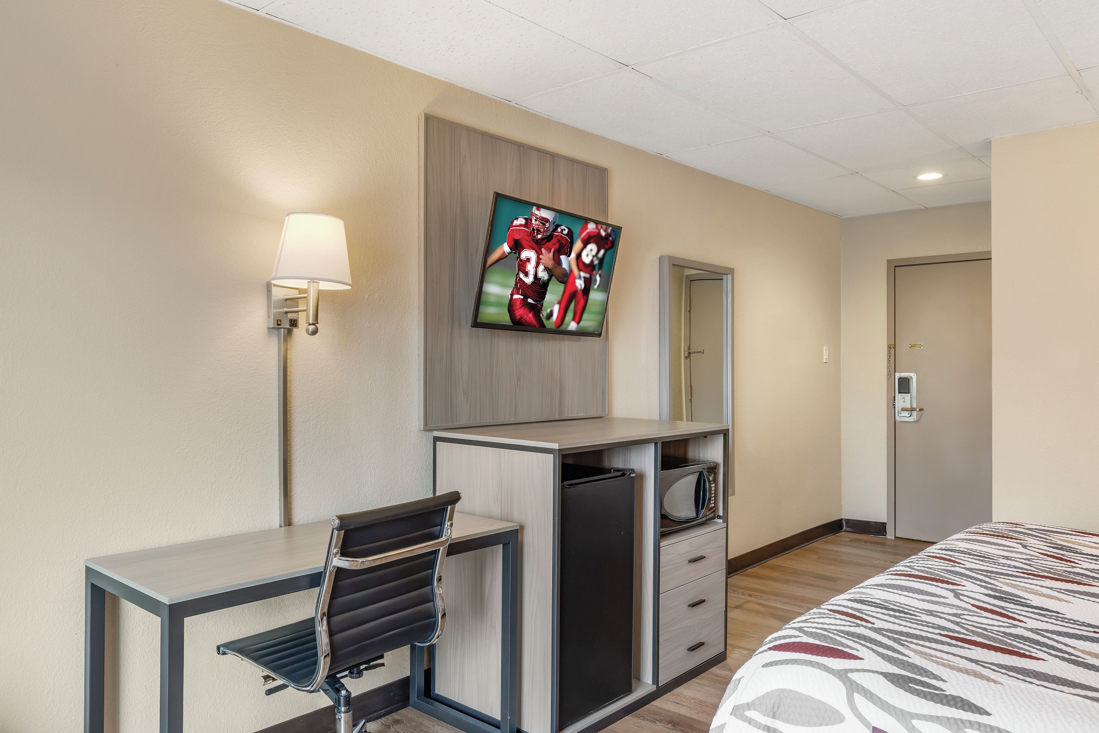 A hotel room with a bed, a desk with a chair, a television mounted on the wall, a microwave, and a door with a lock.