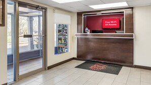 A reception area with a 'Free Wi-Fi All Rooms' sign, a reception desk, a glass door entrance, and a small window with a view. - Red Roof Inn Stroudsburg (Stroudsburg)