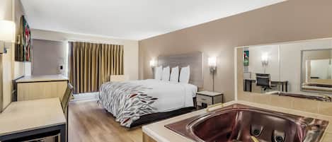 A hotel room with a bed, a desk, a chair, a large mirror, a bathroom with a bathtub, and a shower.